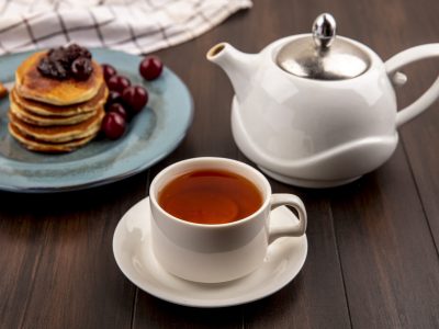 side view of breakfast set with pancake and cherries and fork in plate on plaid cloth and cup of tea with teapot on wooden background